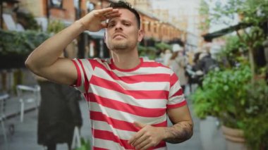 Şehir terasında dövmeli çizgili tişört giyen, şehir atmosferinde bulanık insanlar, bitkiler, sandalyeler ve arka planda binalar olan bir adam selamlıyor..