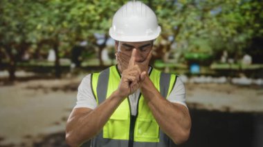 Neon güvenlik yeleği giyen adam mühendis ve beyaz şapka ormanda sessizlik için parmakları dudaklara işaret ediyor..