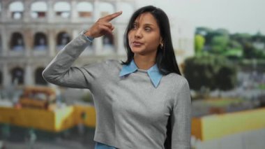 Roma Kolezyumu 'nun yakınlarında, gri bir kazak ve mavi yaka giyen, sembolik bir geçmişi olan İspanyol özelliklerini vurgulayan, ciddi bir ifadeyle jest yapan genç bir kadın..