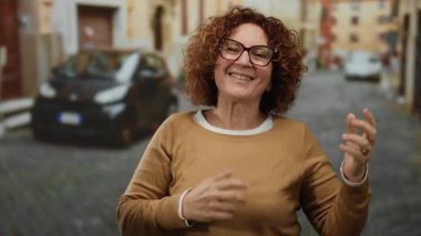 Güneşli bir caddede gitar çalan, gülümseyen ve el kol hareketi yapan bir kadın. Arka planda bulanık arabalar ve tarihi binalar var. Neşeli şehir atmosferini yakalıyor..