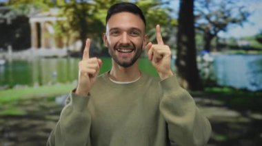 Yeşil bir parkta ağaçlar ve gölle gülümseyen genç bir adam. Üstü açık bir süveter giyip, güneşli günün tadını çıkarıyor..