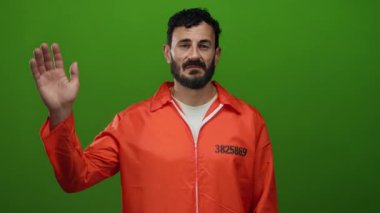 Turuncu hapishane tulumlu İspanyol adam yeşil arka plana karşı duruyor, kasvetli bir ifadeyle elini kaldırıyor, hapis ve kimlik temalarını yansıtıyor..