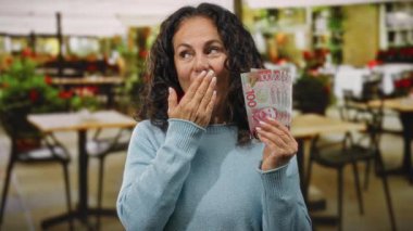 Orta yaşlı bir kadın, yeşillik ve refahı yansıtan açık hava restoranlarının terasında sürpriz bir ifadeyle yeni Zelanda doları tutuyor..