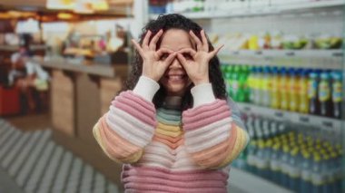 Kapalı alanda gökkuşağı süveteriyle gülümseyen bir kadın canlı bir süpermarkette elleri ve renkli rafları olan parlak ışıklarla gözlük hareketi yapıyor..