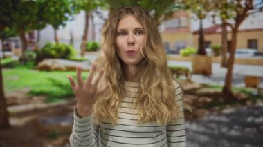 Parkta konuşurken elini kaldıran bir kadın, sarışın, genç, açık havada gülümseyen bir jesti gündelik tarzıyla gösteriyor..