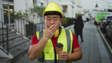 Şehir sokağında elinde kahve fincanı tutarken miğferi ve yeleği olan genç bir adam ağzını kapatıyor..
