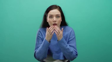 Şaşkınlık ve şaşkınlık ifade eden bir kadın canlı yeşil bir arka plana karşı, mavi bir kazak giyen, esmer oyuncu ve meraklı duyguları vurgulayan, geniş gözlü, eller kaldırılmış.