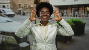 Beyaz ceketle gülümseyen bir kadın şehrin arka planında jestler yaparak öğleden sonraki şehir sahnesinde neşe ve kendine güveni somutlaştırıyor..