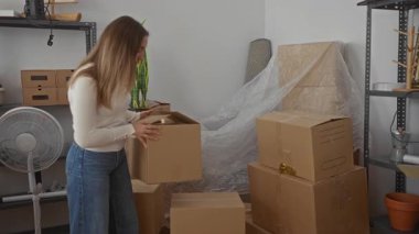 Kadın yeni evinde karton kutuları açıyor. Kollarını açık bir iç mekan ve köşede bir vantilatör ile birleştirerek kollarını kavuşturuyor..