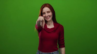 Canlı yeşil bir arka plana gülümseyen, kırmızı bir kazak giyen ve kameraya kendinden emin bir ifadeyle bakan bir kadın..