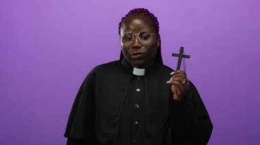 Canlı mor arka planda haç tutan kadın rahip, dinsel kıyafetlerle güvenle gülümsüyor.