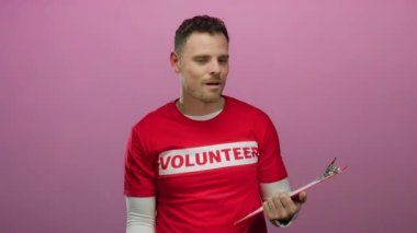 Elinde kırmızı gönüllü gömleği olan genç adam pembe arka planda duruyor. Heyecanlı bir ifadeyle gönüllülük ve coşku gösterisi yapıyor.