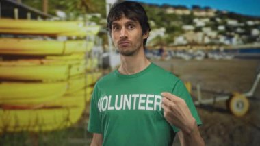 Gönüllü gömlekli bir adam, güneşli gün ortası sahilinde sarı kanolara el hareketi yapar; teşvik.