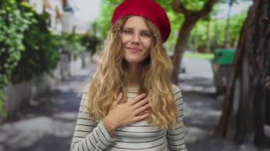 Sarışın bereli ve gözlüklü kadın, yürekten bir gülümseme ile dış görünüşünü aksanlı bir şekilde yansıtırken, kadın şükran dolu bir sokakta ellerini göğsüne koyar..