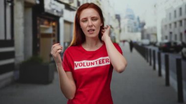 Gönüllü tişörtlü kızıl saçlı bir kadın şehir caddesinde duruyor. Duygularını ifade ediyor. Etrafı şehir mimarisiyle çevrili..