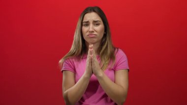 Pembe gömlekli bir kadın, izole edilmiş kırmızı bir arka plana karşı el ele yalvarıyor. Odaklanmış ve derin bir ifadeyle umut, duygu ve samimiyet ifade ediyor..