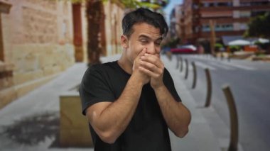 Şehrin sokaklarında ağzını kapatan İspanyol genç adam güneşli bir günde açık şehir ortamında bulanık binalar ve canlı bir atmosferle şaşkınlığını dile getiriyor..