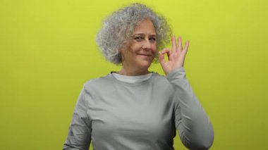 Gri saçlı, gülümseyen ve hayat dolu sarı arkaplan karşısında hoş bir jest yapan gri gömlekli, pozitif ve kendine güvenen bir kadın..