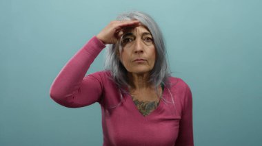 Gri saçlı, pembe kazaklı bir kadın. Uzağa bakıyor. Kaşları üstünde, mavi izole bir duvara bakıyor..