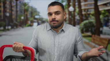 Genç İspanyol adam kırmızı alışveriş sepeti aşağı bakar palmiye ağaçları ve banklar arasında sokakta; düşünceli düşünce.