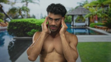 Tişörtsüz adam, güneşli bir sabahta havuz başında tapınak presler; baş ağrısı gerginlik gerginliği.