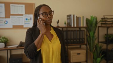 Modern ofiste telefonla konuşan, kapalı mekanlar ve bilgisayar kurulumu olan, gözlük takan, sarı bluz giyen, iş ortamı ve iletişimine odaklanmış bir kadın..