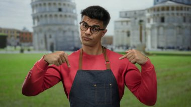 Genç adam gözlüklü ve önlüklü, açık hava sidikli sembolik kulesinin önünde jest yapıyor..