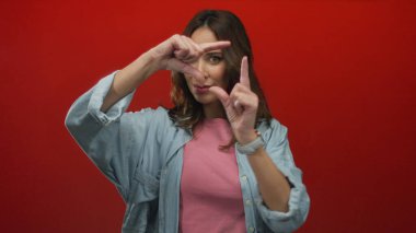 Kadın canlı kırmızı arka plan önünde elleri ile çerçeve yaratıyor günlük kot ve pembe giysiler giyiyor, yaratıcılık yayıyor ve özgüvenli, düşünceli bir ifadeyle odaklanıyor.