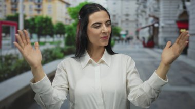 Şehir merkezinde, sokak ortasında el kol hareketi yapan bir kadın neşeli bir ifade ve gün ışığında doğal güzellik, gençlik enerjisi ve özgüvenini simgeler..