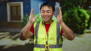 Sarı yelekli bir adam arka planda ağaçlar ve binalar olan şehir sokağında gülümsüyor ve Asyalı yetişkin bir erkeğin yüksek görüş mesafesinde olduğu açık hava sahnesini çekiyor..