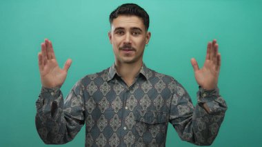 Genç İspanyol adam yeşil arka plan duvarının önünde elleri ile büyük jest yapıyor, desenli gömlek ve küpeler giyiyor, boyunu veya mesafesini ifade ediyor..