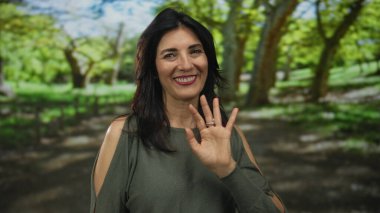 Orta yaşlı bir kadın, güneşli bir ormanda çıplak el sallayarak sıcak bir şekilde gülümserken, sıcak ve samimi bir şekilde selamlıyor..