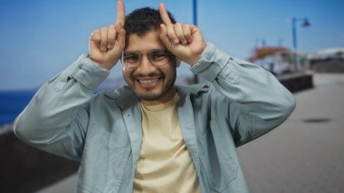 Gümüş gözlüklü adam geniş bir gülümseme yapar ve meşgul sahil caddesinde parmak boynuzları jest; neşe.