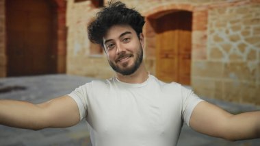 Sakallı İspanyol bir adam eski bir üniversite binasının arka planında kolları ve göğsü açık poz veriyor..