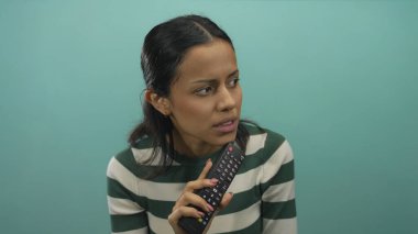 İzole edilmiş yeşil duvar arka planına karşı televizyon kumandası tutan kadın odaklanmış ve düşünceli bir şekilde çizgili gömlek giyerek modern medya yaşam tarzını temsil ediyor.