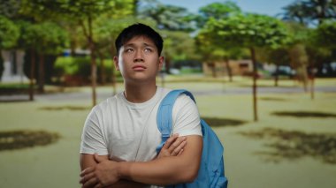 Genç adam güneşli bir parkta kollarını kavuşturmuş, tişört ve sırt çantası giymiş, açık havada bir doğal ortamda iç gözlem anı yakalıyor..