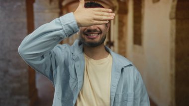 Latin kökenli genç adam, güneş ışığıyla aydınlatılmış tarihi üniversite binasında elleri ile gözlerini kapatırken gülümsüyor; gizlilik gizemi merakı.