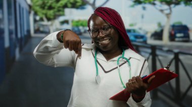 Kadın doktor elinde bir not panosuyla şehir sokaklarında gülümsüyor ve boynunda steteskopla neşeli ve olumlu tavırları vurguluyor.