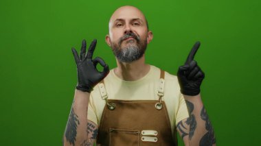 Man with beard and tattoos wearing apron and black gloves gestures ok against green wall background