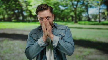 Güneşli bir park ortamında gülümseyen sakallı genç adam, sıradan bir kot ceket giyiyor, açık havada mutluluğu ve rahatlığı ifade ediyor yemyeşil ve ağaçlarla çevrili..
