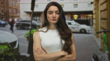 Çapraz kollu beyaz tişörtlü genç İspanyol kadın park etmiş arabalarla kalabalık bir şehir caddesinde duruyor..