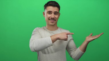 Genç İspanyol bir adam, izole edilmiş yeşil bir arka planda, açık avucunu işaret ederek önerilerde bulunuyor ve dostça bir ifadeyle fikirleri sunuyor..