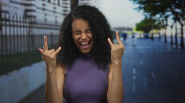 Metal çitli bir şehir caddesinde taş boynuzu hareketi yapan genç İspanyol kadın; rock eğlence partisi enerjisi.