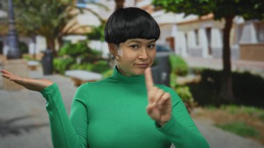 Yeşil tişörtlü genç Çinli kadın barışçıl bir açık hava parkında el kol hareketi yaparak doğayı ve şehir manzarasını harmanlıyor ve sakin bir şekilde reddediliyor..