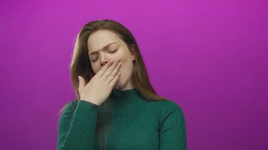 Yeşil kazaklı genç kadın başını eğiyor ve elleri mor stüdyo ortamında ağzını kapatıyor..