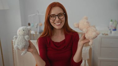 Kızıl saçlı bir kadın rahat yatak odasında sevimli pelüş oyuncaklar tutuyor. Arka planda bir beşiği ve şifoniyeri var. Ev içinde oyuncu bir sahne sergiliyor..