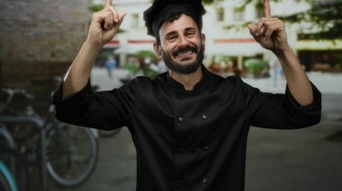 Sokak ortasında gülümseyen sakallı İspanyol erkek şef, gelişigüzel bir şehir ortamında neşeyle işaret ediyor..