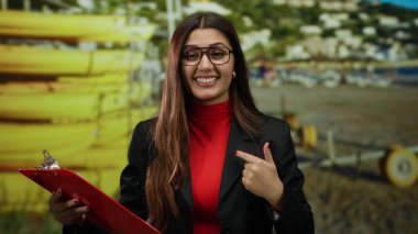 Genç İspanyol kadın deniz kıyısındaki bir yeri işaret ediyor. Elinde açık mavi gökyüzü ve uzak evleri olan güneşli bir arka planda kırmızı bir pano var..