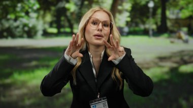 Parktaki kadın, güneşli bir günde arka planda yemyeşil ağaçların arasından süzülen güneş ışığıyla doğal ortamda gözlük ve kimlik kartı takarak anlamlı jestler yapıyor..