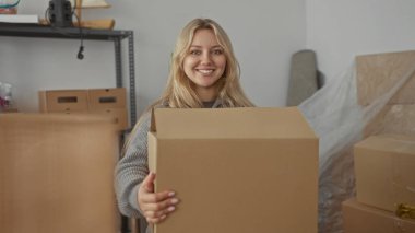 Yeni apartman dairesinde kutuları açan kadın, elinde karton paketle gülümsüyor ve içeriyi düzenliyor..